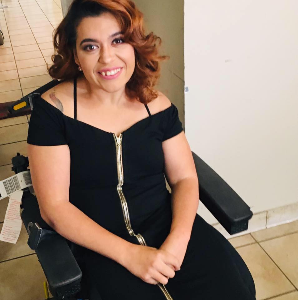 a tanned woman sitting with shoulder length curled reddish brown hair and brown eyes in a wheelchair smiling into the camera with her head tilted wearing a black off the shoulders dress with a gold zipper going down the middle.