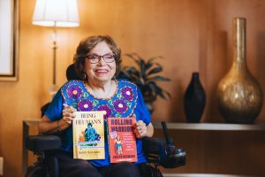 Judy Heumann Smiling, sitting in electric wheelchair holding two books she authored.