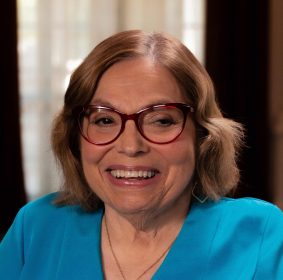 A headshot of Judy Heumann, a white woman with shoulder-length brown hair wearing red glasses, a blue v-neck shirt, and a gold necklace. She is smiling warmly.
