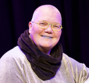 A White queer cis disabled woman of size with a bald head, black lipstick, yellow glasses, velvet top and scarf, and a broad smile.